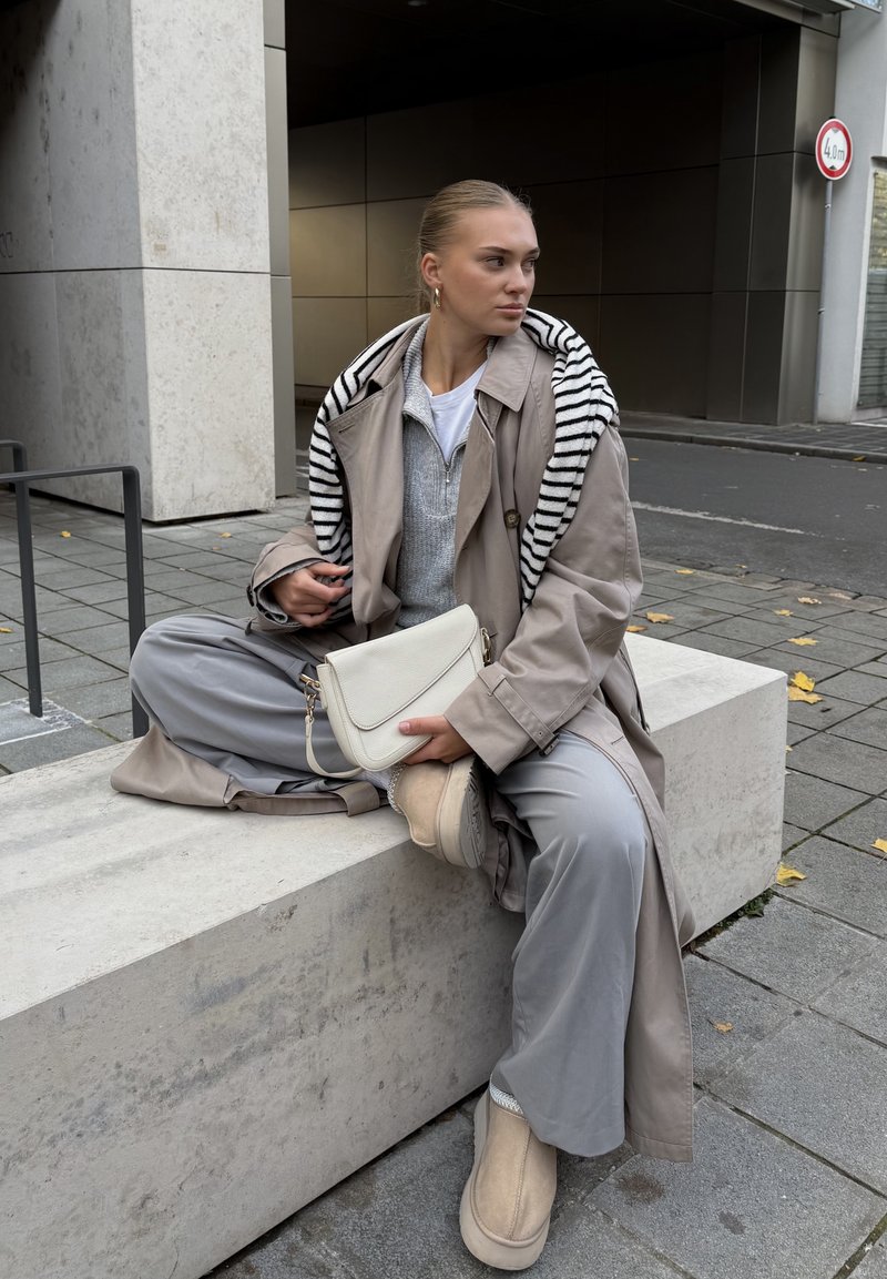 Jovem mulher vestindo um casaco trench coat bege e um suéter às riscas, sentada de pernas cruzadas num banco de betão, segurando uma mala branca, olhando para a direita.