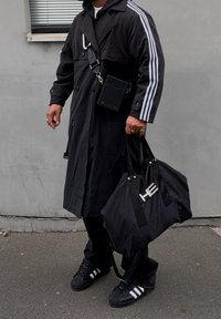Trench-coat noir avec des rayures blanches, sac bandoulière noir et sac de sport. Porte des baskets Adidas noires. Cadre urbain, fond gris.