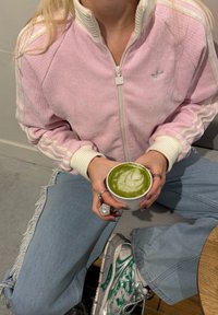 Personne portant une veste Adidas rose et un jean bleu clair, tenant une tasse blanche avec un latte matcha vert, assise avec une jambe croisée.
