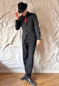 Svart kostym med röd slips, bärande en svart fedora. Kostymen har en skräddarsydd passform, knäppning fram, och en slät textur. Bakgrund av trägolv.