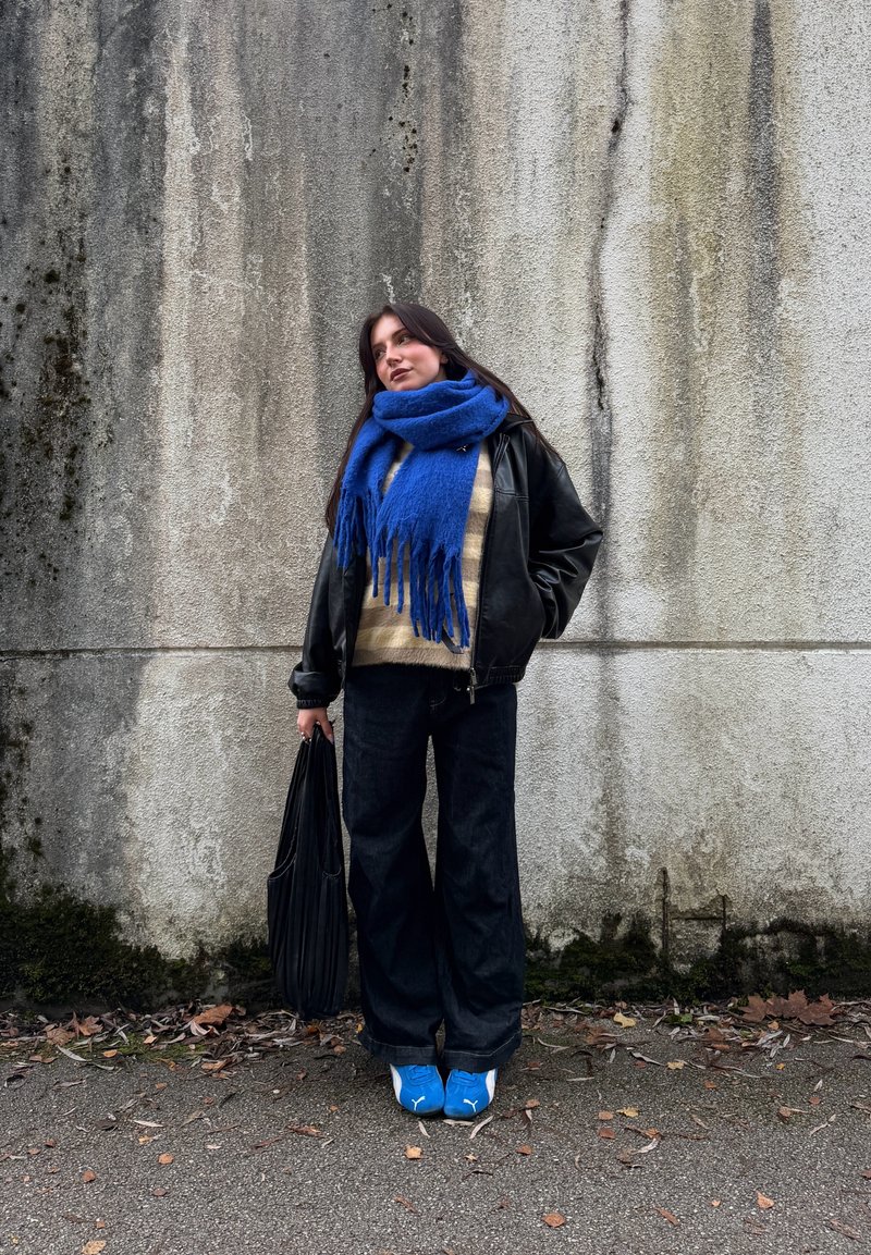 Casaco de couro preto, cachecol de lã azul com franjas, camisola riscada em bege e creme, calças largas de ganga escura, sapatilhas azuis, a segurar uma mala preta.
