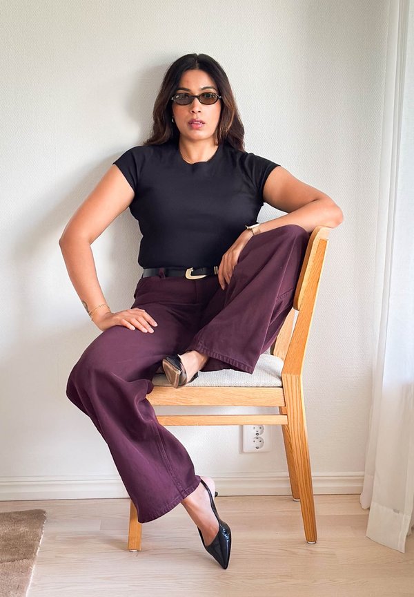Woman wearing dark sunglasses, black shirt, and purple pants sitting on a wooden chair with one leg resting on the seat.