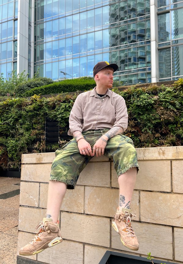 Una persona está sentada en un muro de piedra, vestida con una camisa de cuadros marrón, pantalones cortos de camuflaje y zapatillas beige con toques naranjas. Tatuajes visibles en la pierna.