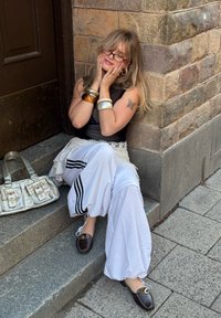 Fekete ujjatlan felső, fehér réteges nadrág fekete csíkokkal, barna slip-on cipők, ezüst kézitáska, karkötők, kőlépcsőkön ül.