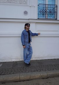 Denim jacket and trousers in light blue, black shirt, black beret, and sunglasses. Leaning against a white wall with decorative accents.