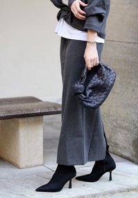 Personne portant un pantalon gris à jambes larges et des bottes à talons noires, tenant un sac à main noir tressé près d'un banc en pierre et d'un mur.