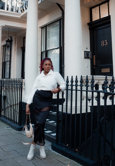 Suéter blanco con cuello, falda negra con detalles de encaje, botines crema y un bolso beige. Se encuentra cerca de una barandilla de hierro forjado negra.