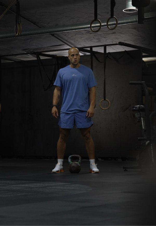 Homme en tenue de sport bleue se tient dans une salle de sport près d'une kettlebell avec des anneaux de gymnastique et un vélo d'exercice en arrière-plan.