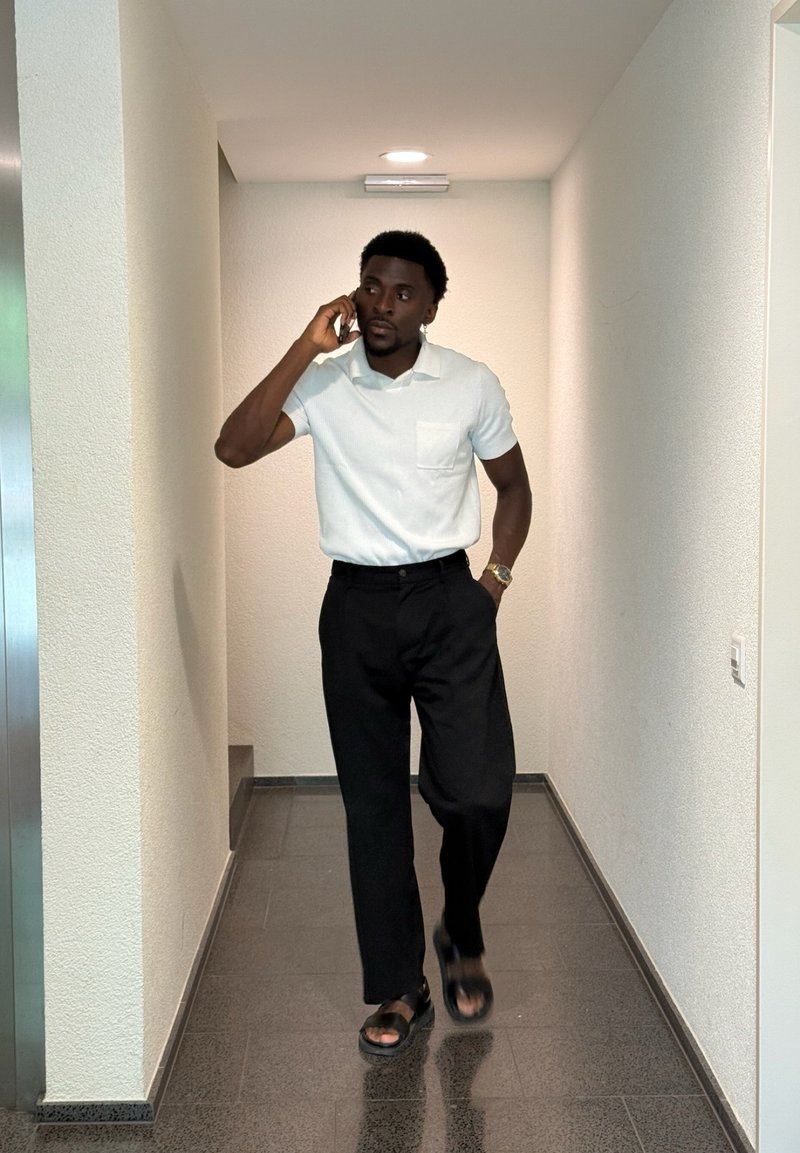 Chemise polo blanche à manches courtes, pantalon noir et sandales noires ; marchant dans un couloir avec des murs texturés et un sol carrelé.