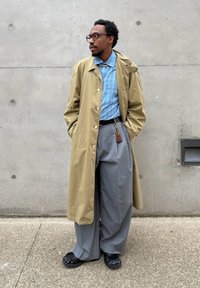 Άνδρας με γυαλιά που φοράει μπεζ παλτό τύπου trench, μπλε πουκάμισο με καρό σχέδιο, γκρι φαρδιά παντελόνια, μαύρα παπούτσια, και στέκεται δίπλα σε τοίχο από σκυρόδεμα.