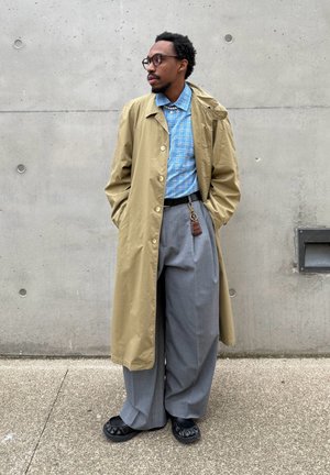 Hombre con gafas vestido con un abrigo beige, camisa a cuadros azul, pantalones anchos grises, zapatos negros, y posando frente a una pared de hormigón.