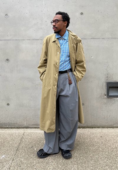 Hombre con gafas vestido con un abrigo beige, camisa a cuadros azul, pantalones anchos grises, zapatos negros, y posando frente a una pared de hormigón.