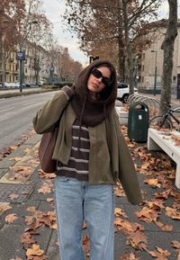 Veste courte verte, capuche en tricot marron, chemise rayée marron et blanche, jeans bleu clair, lunettes de soleil foncées, sac à main en cuir, feuilles d'automne au sol.