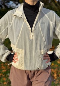 Lekka, kremowa kurtka na zamek z wysokim kołnierzem, teksturowaną tkaniną i przednimi kieszeniami; w połączeniu z brązowymi sportowymi spodniami.