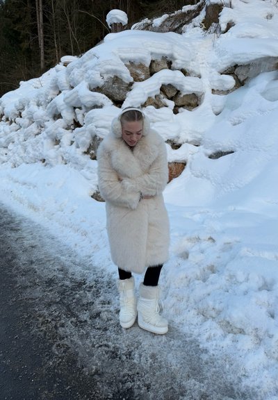 Abrigo de pelaje sintético en color crema, largo y esponjoso, combinado con pantalones negros y botas de nieve blancas. La escena presenta un fondo nevado y un terreno rocoso.