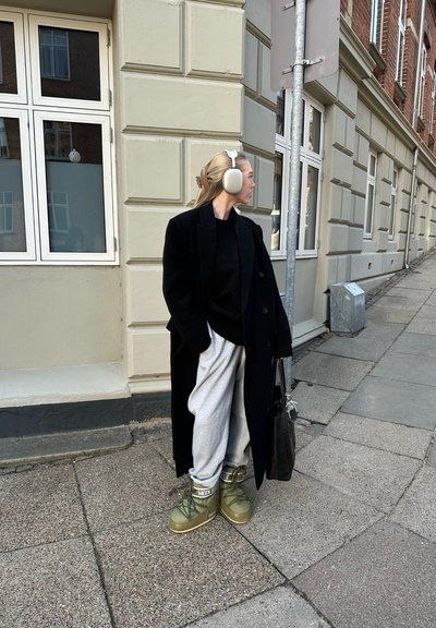 Abrigo de lana negro, suéter negro, pantalones de chándal grises y botas verdes con cordones. También incluye orejeras plateadas y un bolso negro.