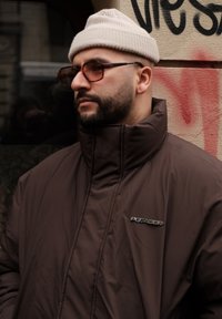 Καφέ επενδυμένο μπουφάν με ψηλό γιακά και λεπτομέρεια λογότυπου, φορεμένο με ανοιχτό μπεζ πλεκτό καπέλο και σκούρα γυαλιά σε μια υφασματική φόντο.