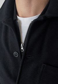 Veste en laine noire avec une fermeture solide à glissière argentée et des boutons, présentant une texture lisse et un détail de poche poitrine. Chemise blanche en dessous.