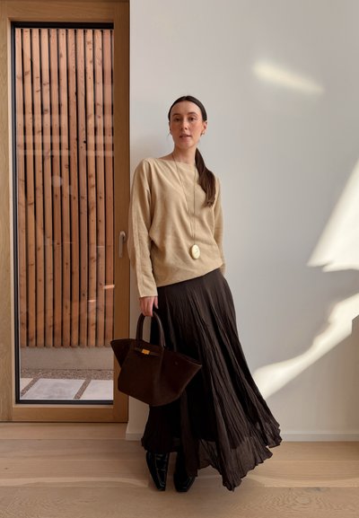 Mujer con blusa beige de manga larga, falda negra plisada, zapatos negros, sosteniendo un bolso marrón oscuro, de pie dentro de un edificio cerca de una puerta de madera y una pared blanca.