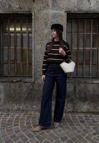 Jeune femme portant un béret noir, un pull rayé, un jean large et des bottes beiges, tenant un sac à main blanc, debout sur une rue pavée.