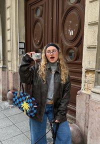 Pruun nahast jakk, hall kõrge kaelusega kampsun, sinised laia säärega teksad ning heegeldatud kott sinise ja musta mustriga ning kollase tähe-detailiga.