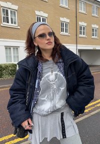 Veste oversize en denim, t-shirt graphique gris avec un motif figuratif, jupe grise à couches avec des accents noirs, et bijoux argentés. Bandeau bleu clair.