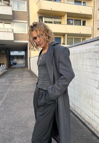 Palton lung, gri închis, peste un top gri mulat, asortat cu pantaloni largi negri. Detaliu cu colier vizibil. Cadru urban cu un perete texturat.