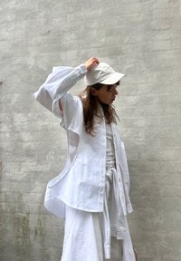 Camisa branca oversized com detalhes recortados nos lados e mangas largas, sobreposta a uma blusa branca simples, complementada por um boné branco.