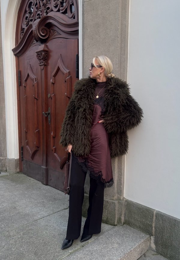 Green fur coat over a brown satin dress with lace trim, paired with black wide-leg pants and pointed black shoes. Dark wooden door background.