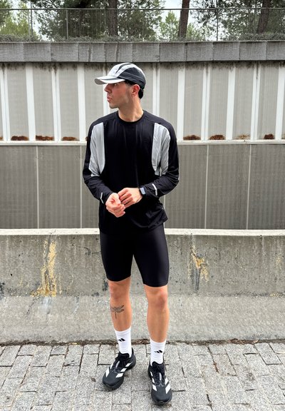 Hombre con ropa deportiva negra y gris, gorra y zapatillas de correr, de pie sobre suelo pavimentado cerca de una barrera de hormigón.
