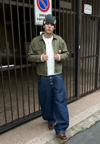 Jeune personne portant un bonnet gris orné d'épingles, une veste olive, un pull blanc, un jean large bleu et des chaussures marron, debout près d'un portail métallique avec des panneaux d'interdiction de stationner.