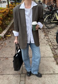 Barna, túlméretezett blézer fehér inggel, világoskék farmerrel és fekete cipővel párosítva. Fekete táskát és zöld kávésbögrét tart.