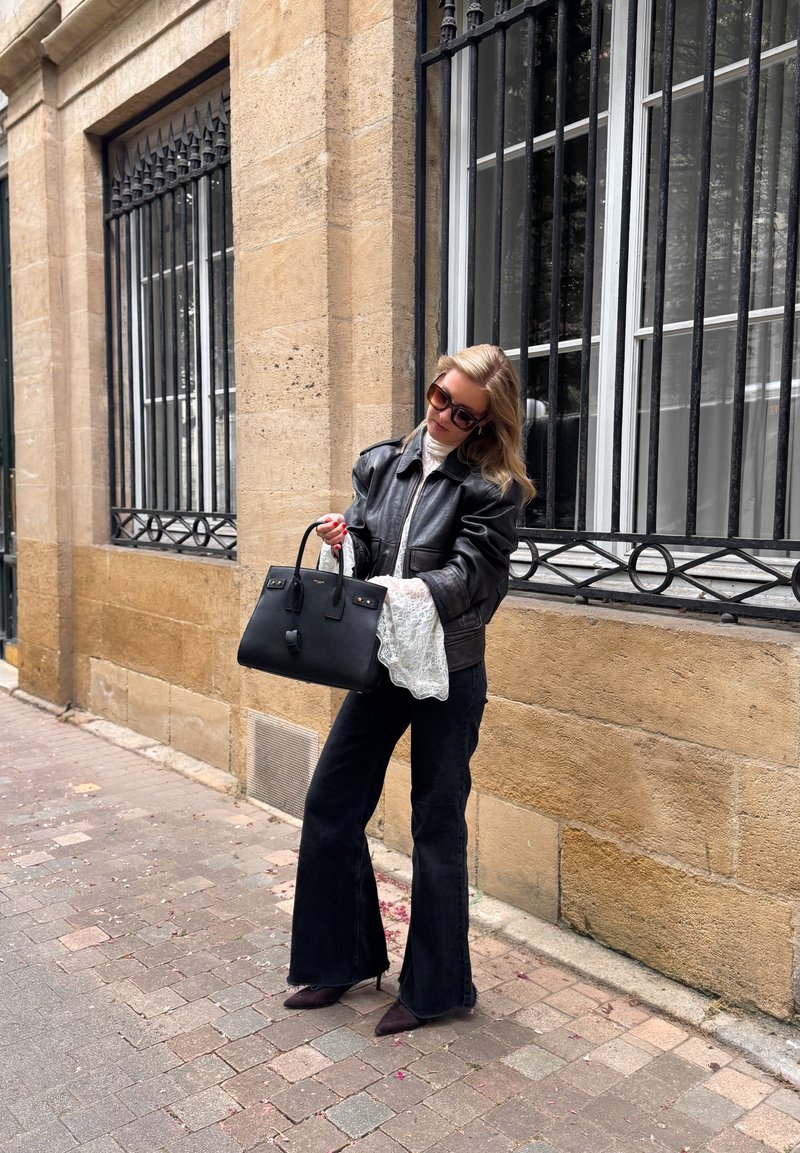 Femme portant une veste en cuir noire, un chemisier en dentelle blanche, un pantalon noir évasé et des lunettes de soleil, tenant un sac à main noir dans une rue pavée.