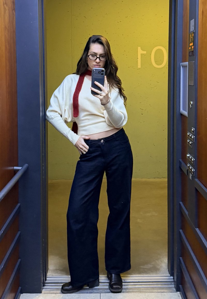 Jeune femme aux longs cheveux et lunettes pose pour un selfie dans un miroir d’ascenseur, portant un pull crème, un pantalon large noir et des bottes noires.