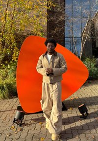 Bēša virsdrēbju komplekts ar pieguļošu jaku un vaļīgām biksēm, kopā ar oranžu abstraktu skulptūru ārējā vidē ar rudens lapām.