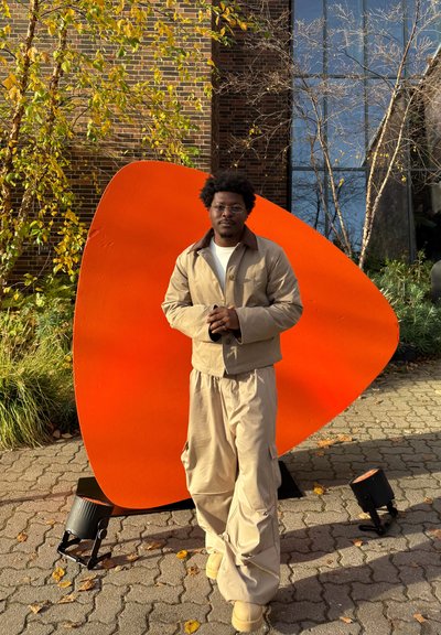 Conjunto de ropa exterior beige con chaqueta entallada y pantalones sueltos, combinado con una escultura abstracta naranja en un entorno al aire libre con follaje otoñal.