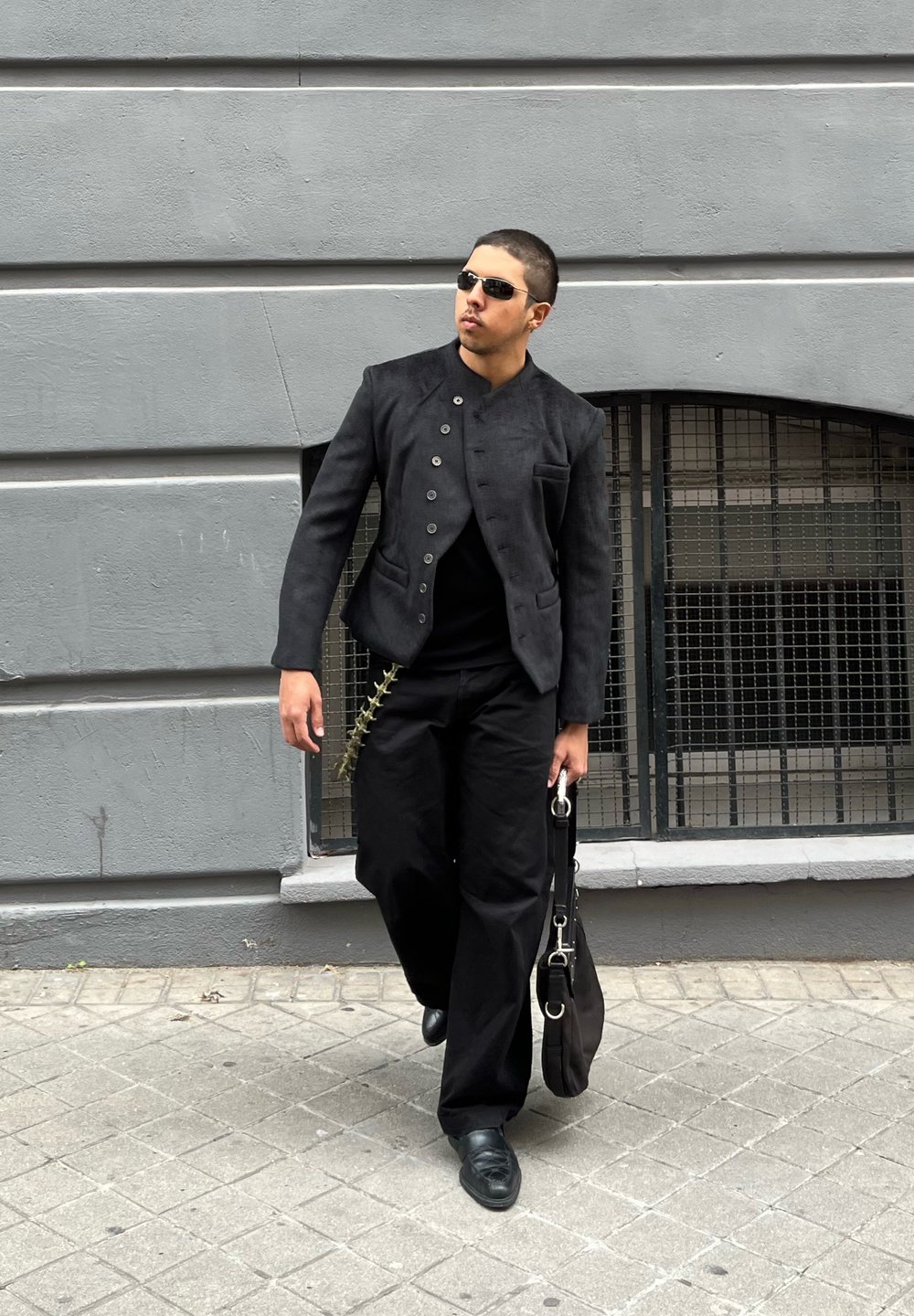 Black textured jacket with a high collar and buttons, paired with loose black trousers. Carrying a black bag featuring a metallic chain detail.