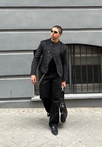 Black textured jacket with a high collar and buttons, paired with loose black trousers. Carrying a black bag featuring a metallic chain detail.