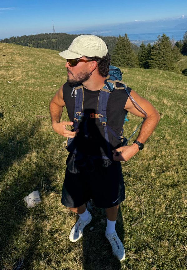 Hombre con gorra, gafas de sol, camiseta negra sin mangas y pantalones cortos ajusta mochila de senderismo en colina cubierta de hierba con árboles y lago al fondo.