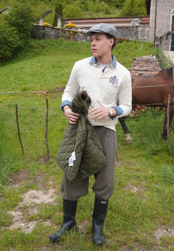 Jeune homme en tenue campagnarde tenant une veste verte, debout sur l'herbe près d'un cheval brun clôturé et d'un bâtiment en pierre dans un environnement rural.