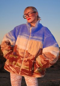 Veste zippée en polaire avec des dégradés de bleu et de beige ; présente un imprimé de scène naturelle, une poche beige et des manches surdimensionnées.