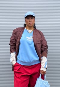 Casaco bomber castanho, sweatshirt azul claro com logótipo, calças vermelhas e boné azul claro. Acessórios incluem corrente dourada e bolsa azul.