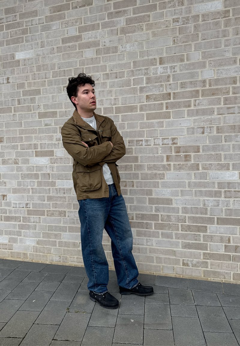Homme debout, les bras croisés, portant une veste marron, une chemise grise, un jean bleu et des chaussures noires devant un mur de briques gris clair.