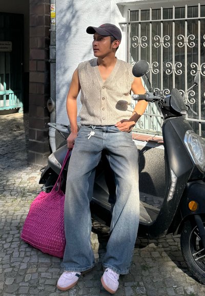 Chaleco de punto gris sin mangas, jeans de pierna ancha azul claro, bolso trenzado rosa y zapatos claros. Scooter negro aparcado al fondo.