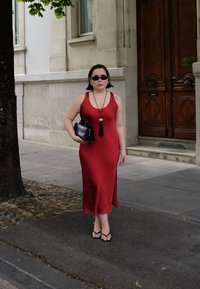 Donna in un vestito rosso senza maniche, sandali neri, occhiali da sole e borsa nera che cammina sul marciapiede della città vicino a un edificio in pietra e una porta di legno.