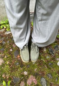 Harmaat mokkakengät pyöreällä muotoilulla, jossa on nauhat ja kumipohja, yhdistettynä löysiin vaaleanharmaisiin housuihin sammalmättäällä.