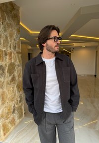 Camisa de botones de color gris oscuro con cuello y dos bolsillos en el pecho, llevada sobre una camiseta blanca. Pantalones gris con rayas finas. Entorno interior moderno con pared de piedra.