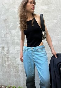 Bodysuit preto sem mangas, jeans azul claro com degradê escuro, mala preta com padrão abstrato branco, segurando uma jaqueta preta.