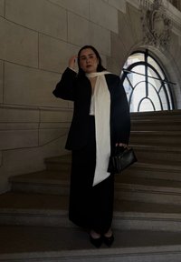 Blazer nero su misura e gonna con foulard bianco. Borsa nera e tacchi a punta. Lo sfondo presenta gradini in pietra e una grande finestra.