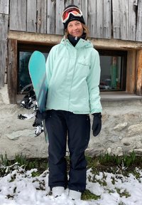 Άτομο που φοράει μιντ πράσινο μπουφάν σνόου και μαύρο παντελόνι σκι, κρατώντας ένα σνόουμπορντ. Στο φόντο φαίνεται ξύλινο καλύβι και χιονισμένο έδαφος.